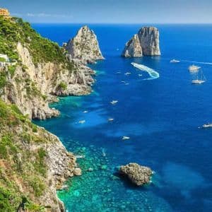 An aerial view of a rugged coastline with large rock formations and several boats sailing on the clear blue sea.