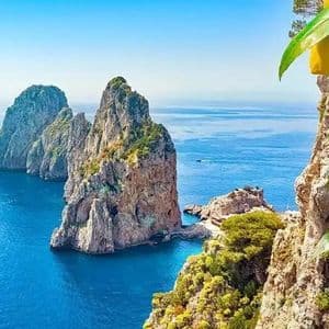 A close-up of lemons on a branch overlooking two large rock formations rising from a bright blue sea under a clear sky.