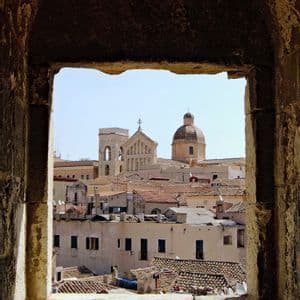 A historic city skyline with a church bell tower and dome, viewed through a rough, stone arched window under a clear sky.