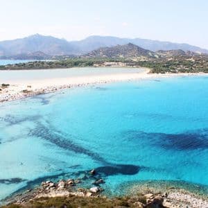 An aerial view of a sandy isthmus beach with turquoise water on both sides, with people swimming and two sailboats floating in the sea.