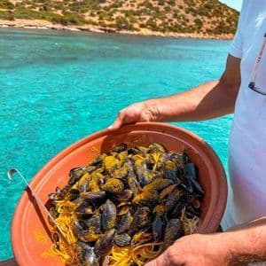 A person holds a large terracotta bowl of spaghetti with mussels, topped with yellow seasoning, over turquoise water.