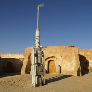 Ein weißer, futuristischer Turm steht inmitten sandfarbener Lehmgebäude in einer Wüstenlandschaft unter klarem blauem Himmel.