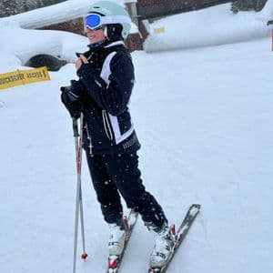 Una persona con casco y gafas de ventisca está de pie sobre unos esquís en una pendiente nevada mientras sostiene bastones de esquí.