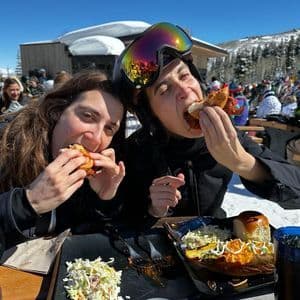 Dos mujeres de un viaje en grupo de WeRoad comen bocadillos en una mesa al aire libre en una estación de esquí soleada y nevada.