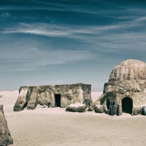 Sandfarbene Gebäude mit Kuppeldach stehen in einer weiten Wüste unter einem gestreiften blauen Himmel.