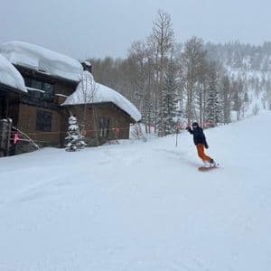 Una persona con chaqueta oscura y pantalones anaranjados hace snowboard en una pendiente nevada junto a casas y árboles cubiertos de nieve.