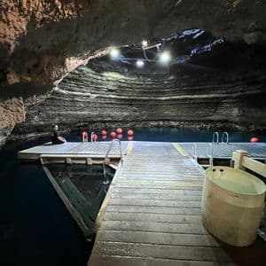 Una persona se sienta en un muelle de madera sobre agua azul oscura dentro de una cueva grande e iluminada.