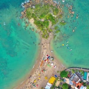 Una vista aerea dall'alto di una lingua di sabbia che collega una città costiera a un isolotto roccioso e verde in acqua turchese, con persone e ombrelloni colorati sulla spiaggia.