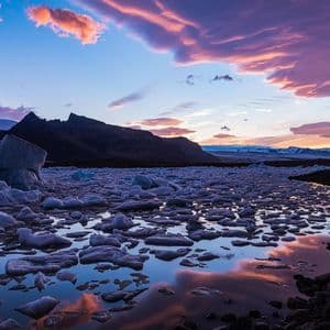 Iceberg galleggiano in un lago glaciale al tramonto con montagne sullo sfondo e nuvole colorate che si riflettono sull'acqua.