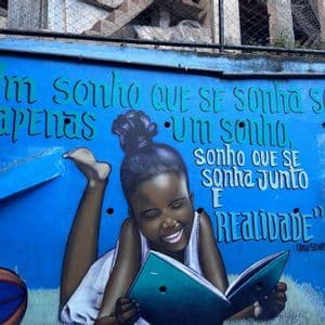 Un murale colorato su una parete blu brillante raffigura una bambina sorridente sdraiata che legge un libro, circondata da una citazione in portoghese.