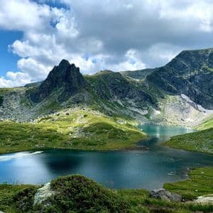 Un lac de montagne aux eaux claires et bleues est niché dans une vallée de montagnes verdoyantes et rocheuses sous un ciel partiellement nuageux.