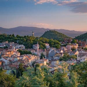 Une ville aux bâtiments colorés et aux toits de tuiles est nichée au milieu de collines verdoyantes sous un ciel de coucher de soleil.