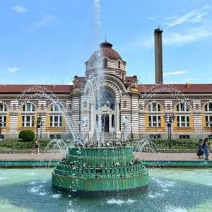 Une fontaine verte à plusieurs niveaux projette de l'eau devant un majestueux bâtiment historique coiffé d'un dôme.
