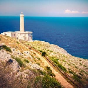A tall white lighthouse on a grassy cliffside overlooking the deep blue sea, with a dirt path in the foreground.