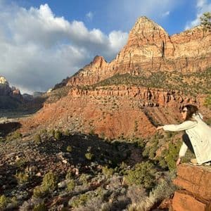 Una mujer con chaqueta blanca se cuclillas en una cornisa de roca con vistas a un vasto cañón de roca roja bajo un cielo parcialmente nublado