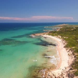 An aerial view of a long, curving coastline with a sandy beach, turquoise water, and lush green vegetation under a clear blue sky.
