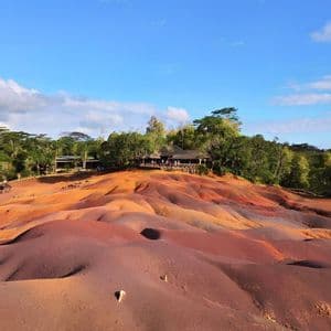 Un paesaggio di dune ondulate e multicolori nei toni del rosso e del viola, con una piattaforma panoramica e una foresta sullo sfondo.