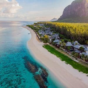 Una vista aerea di un resort su una spiaggia di sabbia bianca, delimitata da acque turchesi e una montagna boscosa sotto un cielo parzialmente nuvoloso.