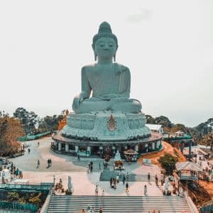 Una vista aerea di una gigantesca statua bianca del Buddha assisa su una collina, con turisti che camminano nella piazza sottostante.
