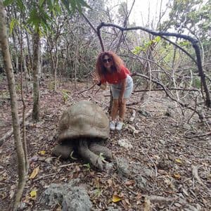 Una donna dai capelli rossi e ricci posa con il segno della pace accanto a una tartaruga gigante su un terreno foglioso in una foresta.
