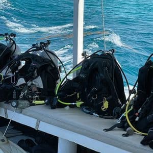 Attrezzatura per immersioni subacquee, incluse bombole e GAV, su una panca di una barca con l'oceano turchese sullo sfondo.