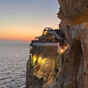 Un ristorante a picco sulla scogliera è illuminato da luci calde che si affacciano sull'oceano al tramonto, visto da una grotta.