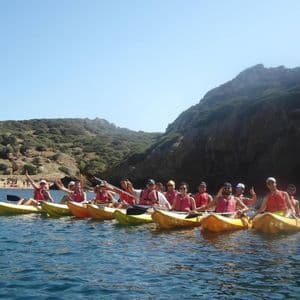 Un gruppo WeRoad in viaggio posa su kayak gialli sul mare, con una costa rocciosa e verde sullo sfondo sotto un cielo sereno.