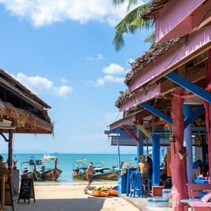 Una vista tra due vivaci chioschi sulla spiaggia, affacciata su un mare turchese con barche a coda lunga ormeggiate sulla sabbia.