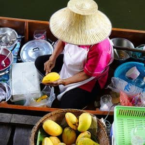 Una vista dall'alto di un venditore con un cappello di paglia che affetta un mango giallo maturo mentre è seduto in una piccola barca.