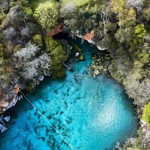 Una veduta aerea di un cenote turchese in una foresta, con una zipline che lo attraversa e persone che nuotano nell'acqua cristallina.