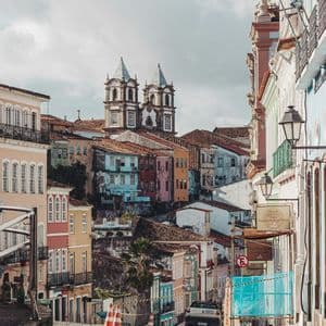 Una vista lungo una ripida strada acciottolata verso colorati edifici storici e una chiesa su una collina sotto un cielo nuvoloso.