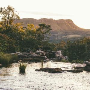 Un fiume scorre su una sporgenza rocciosa in una foresta, con una mesa montuosa visibile sullo sfondo durante il tramonto.