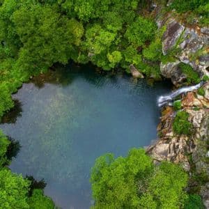 Vista aerea dall'alto di una cascata che si getta in una piscina naturale cristallina, circondata da una lussureggiante foresta verde e rocce.