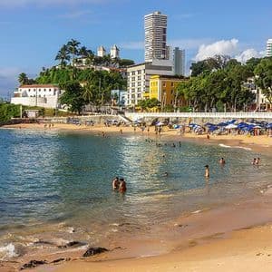 Una spiaggia urbana soleggiata con persone che nuotano nella baia e si rilassano sulla sabbia, con edifici arroccati su una collina verdeggiante sullo sfondo.