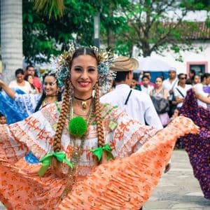 Eine Frau in einem farbenfrohen traditionellen Kleid und Kopfschmuck lächelt, während sie bei einer Freiluftparade tanzt.