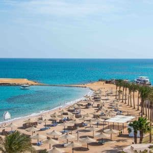 Vista dall'alto: spiaggia sabbiosa, palme, ombrelloni di paglia e mare turchese con isolotto.
