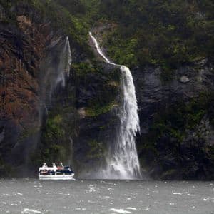 Un'imbarcazione turistica naviga in acque agitate ai piedi di due potenti cascate che si riversano da una scogliera rocciosa e lussureggiante.
