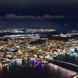 Veduta aerea di una città costiera innevata illuminata di notte, con un ponte illuminato che attraversa l'acqua e montagne sullo sfondo.