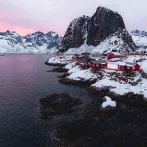 Case di legno rosse si ergono su una costa rocciosa e innevata accanto ad acque scure, con montagne innevate sotto un cielo rosa.