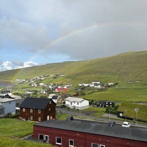 Un arcobaleno si inarca su un piccolo villaggio adagiato ai piedi di una grande collina verde sotto un cielo nuvoloso.
