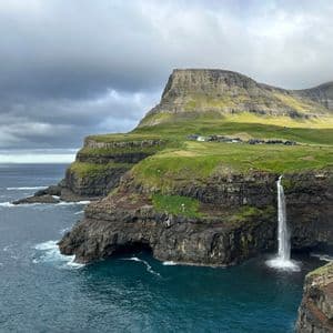 Una cascata si getta da una scogliera verde nell'oceano, con un piccolo villaggio visibile in cima alla scogliera sotto un cielo nuvoloso.