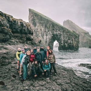 Un gruppo WeRoad in posa su una costa rocciosa, con grandi faraglioni e un arco naturale alle spalle, sotto un cielo nuvoloso.