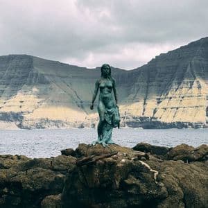 Una statua di bronzo di una selkie che tiene la sua pelle di foca si erge su rocce in riva al mare, di fronte a una scogliera montuosa stratificata.
