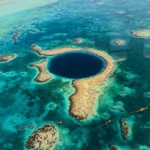 Veduta aerea di un grande buco blu circolare circondato da una barriera corallina in acque oceaniche turchesi, con l'ala di un aereo visibile.