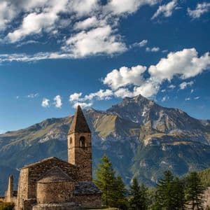 Una rustica chiesa in pietra con campanile si erge su una collina, con una vasta catena montuosa sullo sfondo, sotto un cielo azzurro e nuvoloso.