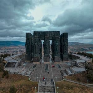 Veduta aerea di un imponente monumento con alte colonne scolpite, mentre persone camminano sulla sua piazza di pietra sotto un cielo nuvoloso.