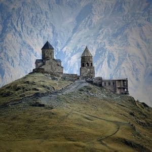 Una chiesa di pietra e un campanile si stagliano su una collina erbosa, con un'imponente e aspra catena montuosa a far da sfondo.