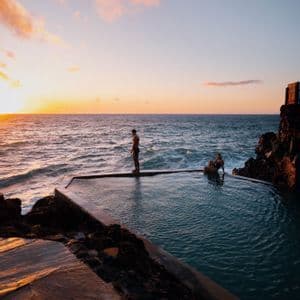 Un uomo in piedi e una donna seduta a bordo di una piscina a sfioro con vista sull'oceano, durante un tramonto colorato.