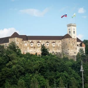 Un gran castillo de piedra con una destacada torre del reloj se alza sobre una exuberante colina arbolada bajo un cielo azul.