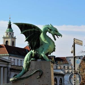 Una estatua de dragón verde con las alas extendidas se asienta sobre un pedestal, con edificios históricos y dos torres de iglesia al fondo.